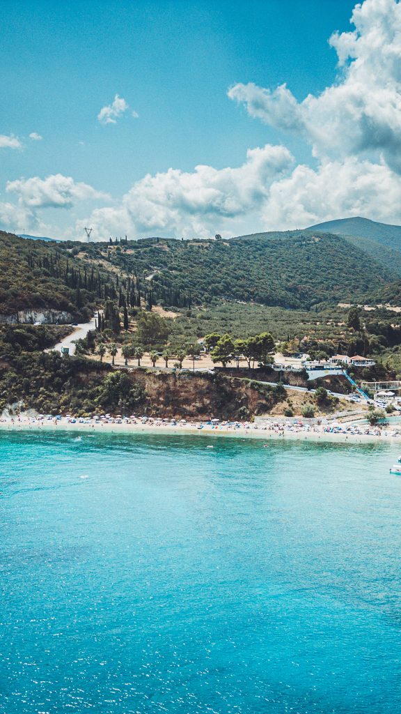 Drohnenaufnahme vom Makris Gialos Beach auf Zakynthos mit Bergen, Zypressen und türkisfarbenem Wasser im Sommerlicht