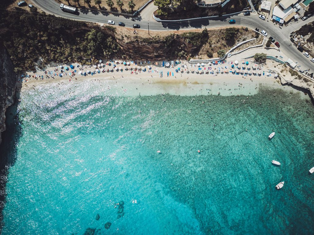 Luftaufnahme vom Makris Gialos Strand auf Zakynthos mit Menschen, bunten Schirmen und glitzerndem Wasser an einem heißen Sommertag