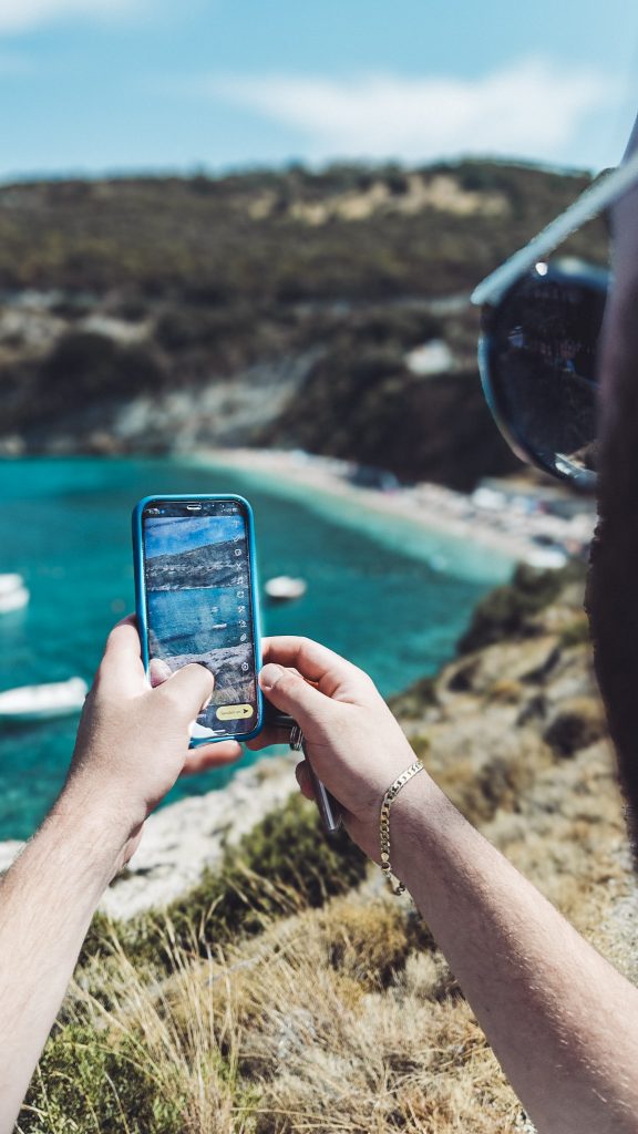 Hände mit Smartphone fotografieren den Strand Makris Gialos auf Zakynthos, Fokus auf das Display mit türkisblauem Wasser im Hintergrund