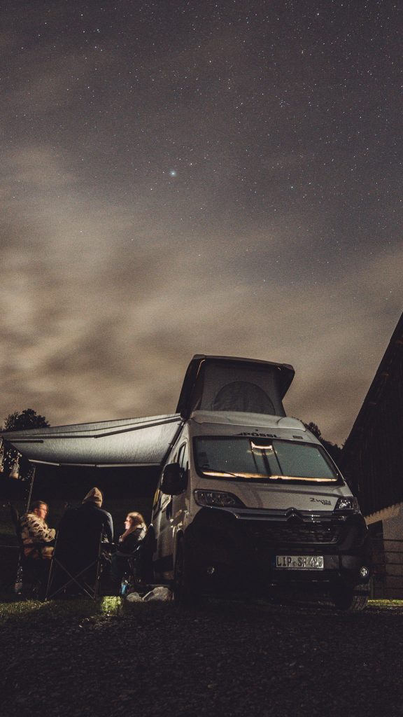 Ein Camper unter einem funkelnden Sternenhimmel – davor eine Gruppe Menschen im Gespräch, warm eingepackt. Dieses Bild fängt die Essenz eines perfekten Campingabends ein: Gemeinschaft, Natur und Freiheit.