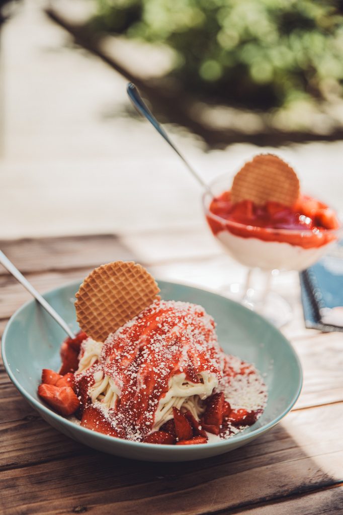 Zwei Eisbecher mit Erdbeeren, Waffel und Löffel auf einem Holztisch mit Natur im Hintergrund