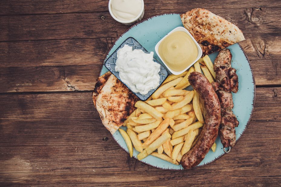 Von oben fotografierter Teller mit Grillwurst, Pommes, Pita und Dips auf rustikalem Holztisch