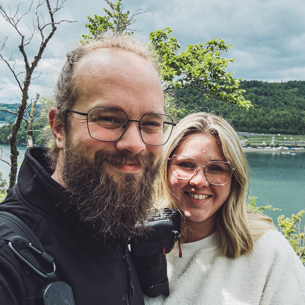 Paar mit Kamera am Seeufer, lächelt während einer Wanderung in die Kamera