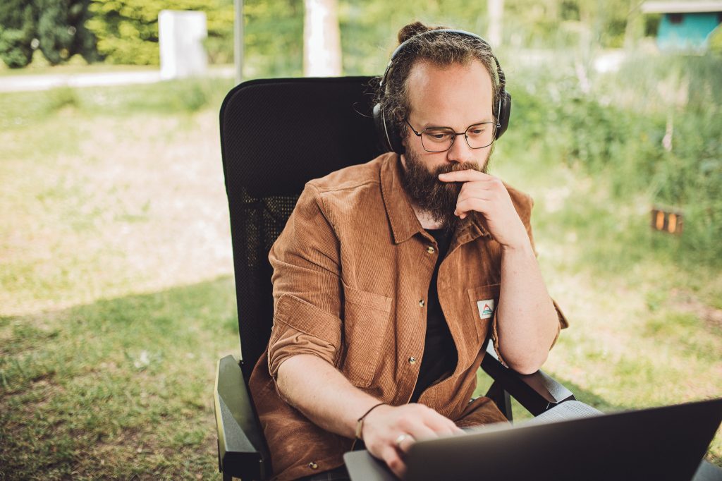 Mann mit Kopfhörern arbeitet am Laptop im Freien auf einem Campingstuhl