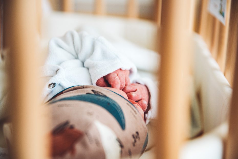 Neugeborenes Emil liegt im Kinderbett in Detmold mit gefalteten Händen, eingerahmt von Holzstäben – ein zarter Moment voller Geborgenheit.