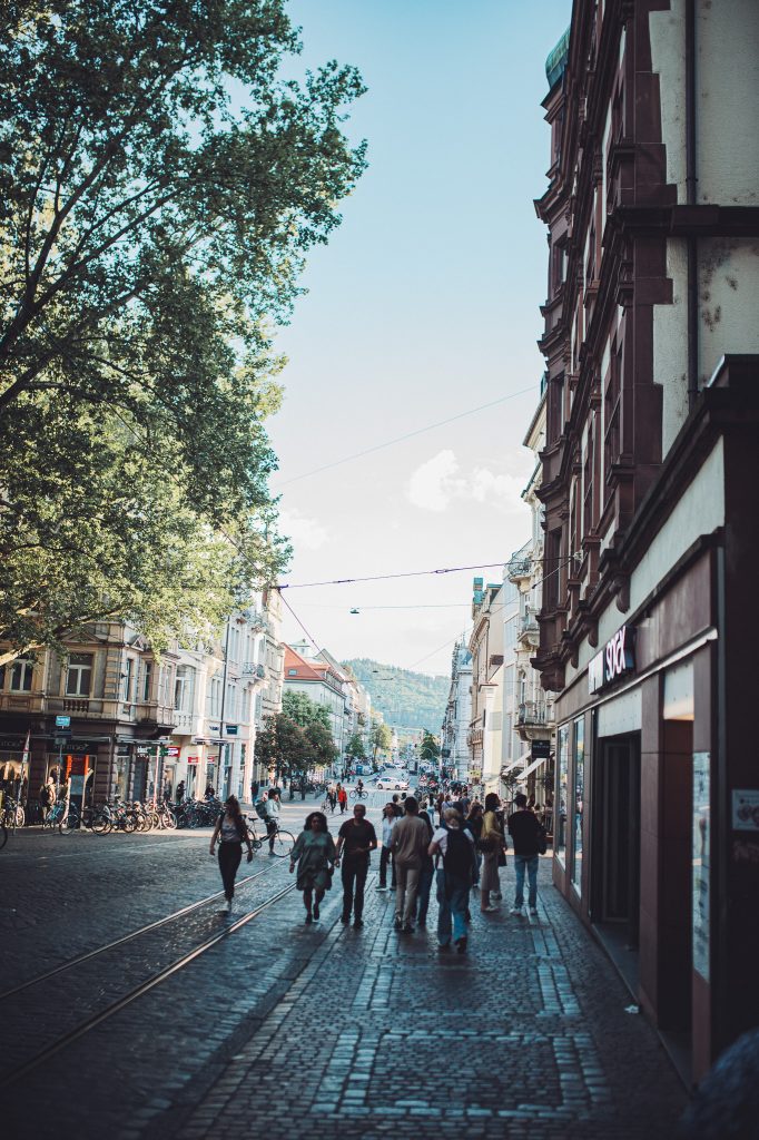 Fußgängerzone in Freiburg mit alten Gebäuden, Geschäften und Menschen bei Sonnenschein