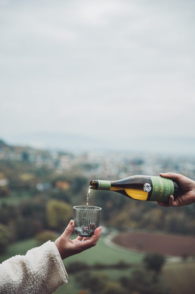 Person gießt Weißwein in ein Glas mit Blick über Wittlich und herbstliche Landschaft im Hintergrund.