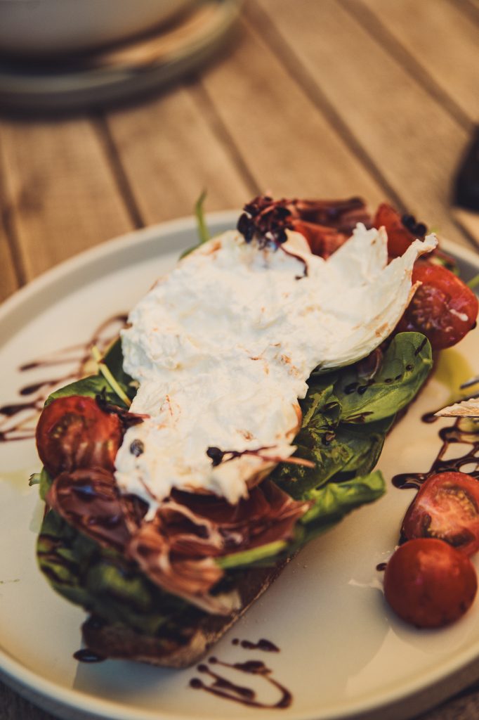 Frisch angerichtetes Burrata-Brot mit Schinken, Spinat und Tomaten auf einem weißen Teller.