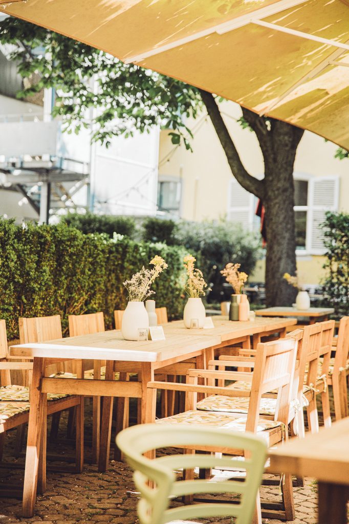 Holztisch mit Stühlen auf der sonnigen Terrasse des Café Marmell in Freiburg, dekoriert mit Vasen und Trockenblumen.