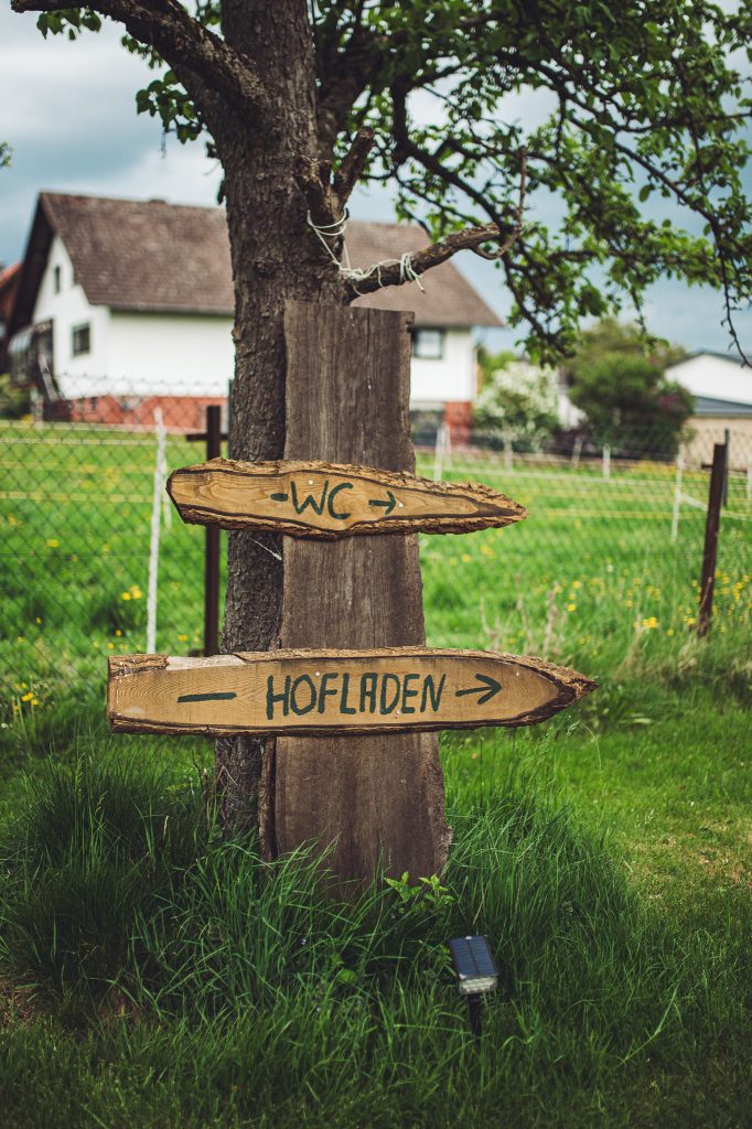 Holzwegweiser an einem Baum mit Hinweisen zu WC und Hofladen auf Kochs Hof in Hessen