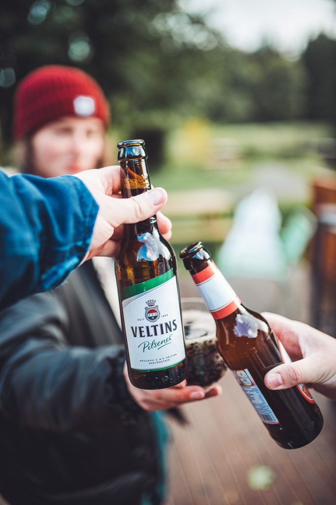 Zwei Personen stoßen mit Bierflaschen im Außenbereich des Campingplatzes an, unscharfer Hintergrund mit Natur