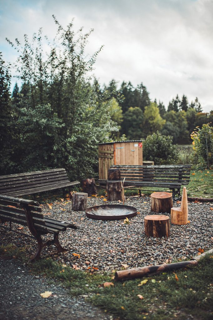 Runde Feuerstelle mit Holzstämmen und Holzbänken auf Kiesfläche umgeben von Bäumen auf dem Campingplatz im Harz