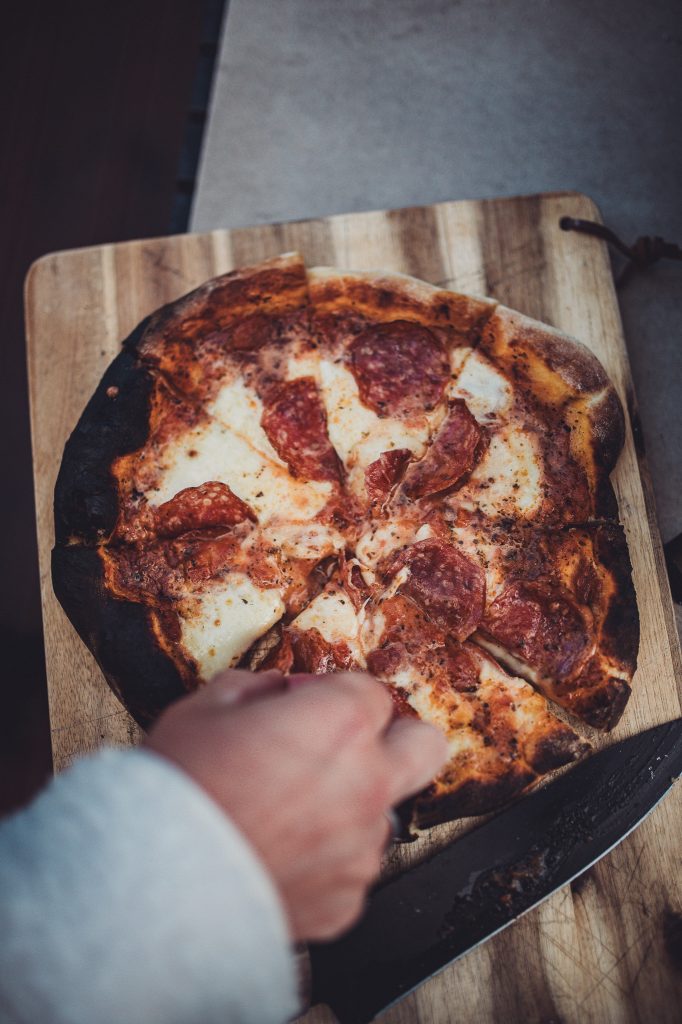 Frisch gebackene Salami Pizza mit angebackenem Rand wird auf einem Holzbrett mit Messer in Stücke geschnitten