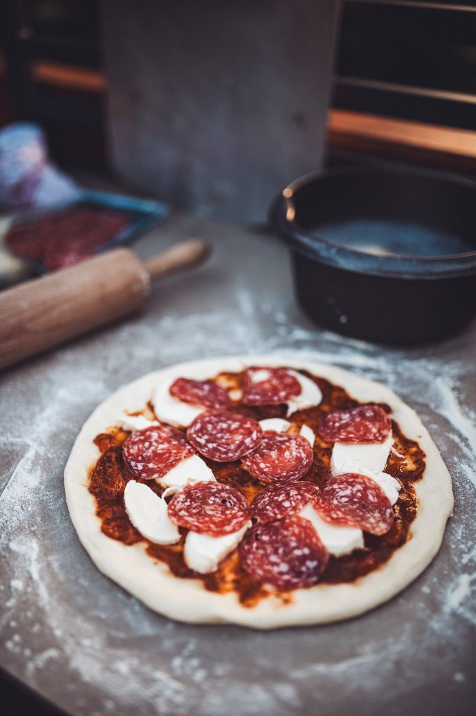 Runde ungebackene Pizza mit Salami und Mozzarella auf bemehlter Arbeitsfläche in der Outdoor Küche des Campingplatzes