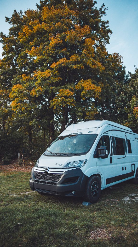 Weißer Campervan auf einem Stellplatz im Harz vor großen herbstlichen Bäumen bei Abendlicht