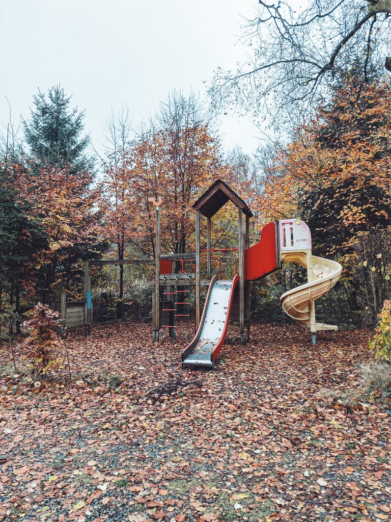Spielplatz mit Rutsche und Kletterelementen im Herbstlaub, umgeben von bunten Bäumen am Camping des Charmilles in Belgien