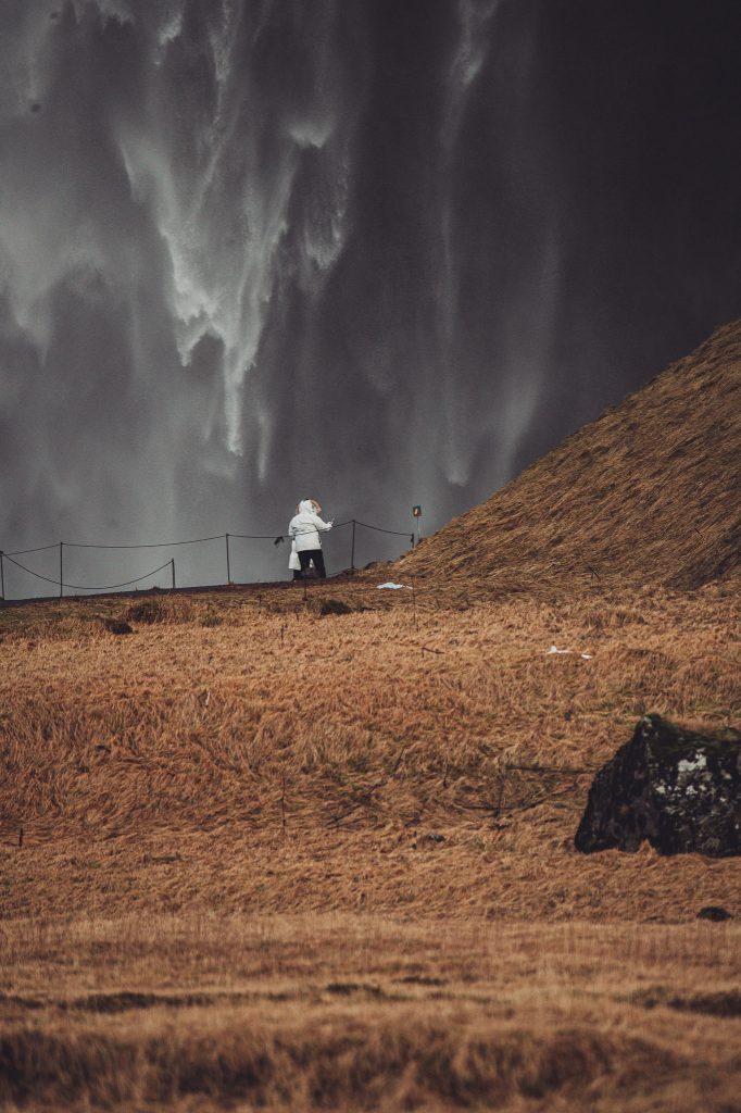 Person in weißer Jacke steht vor der herabstürzenden Wasserwand am Seljalandsfoss in Island