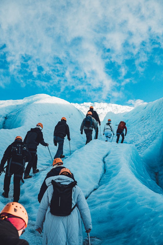 Eine Wandergruppe mit orangenen Helmen und Rucksäcken steigt eine Eisflanke am Gletscher hinauf.