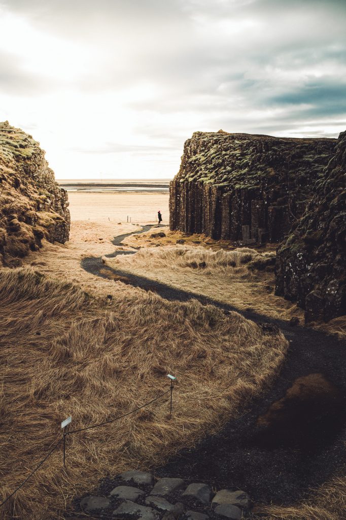 Pfad durch die Basaltformation Dverghamrar in Island mit hohem Fels, hellem Gras und einer Person in der Ferne