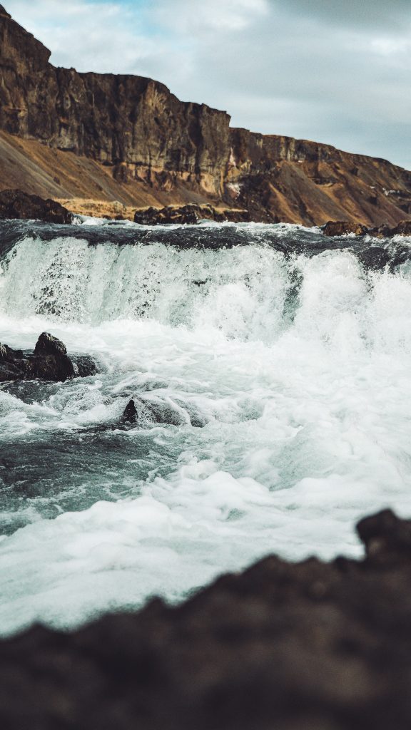 Hochformat vom Fossálar Wasserfall in Island mit weißer Gischt, dunklen Steinen und steiler Felslandschaft im Hintergrund
