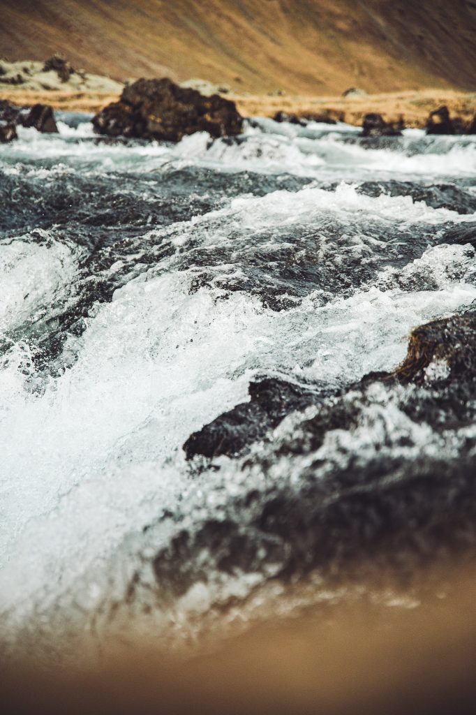 Stromschnellen am Fossálar Wasserfall in Island mit schäumendem Wasser, dunklen Lavasteinen und brauner Landschaft im Hintergrund
