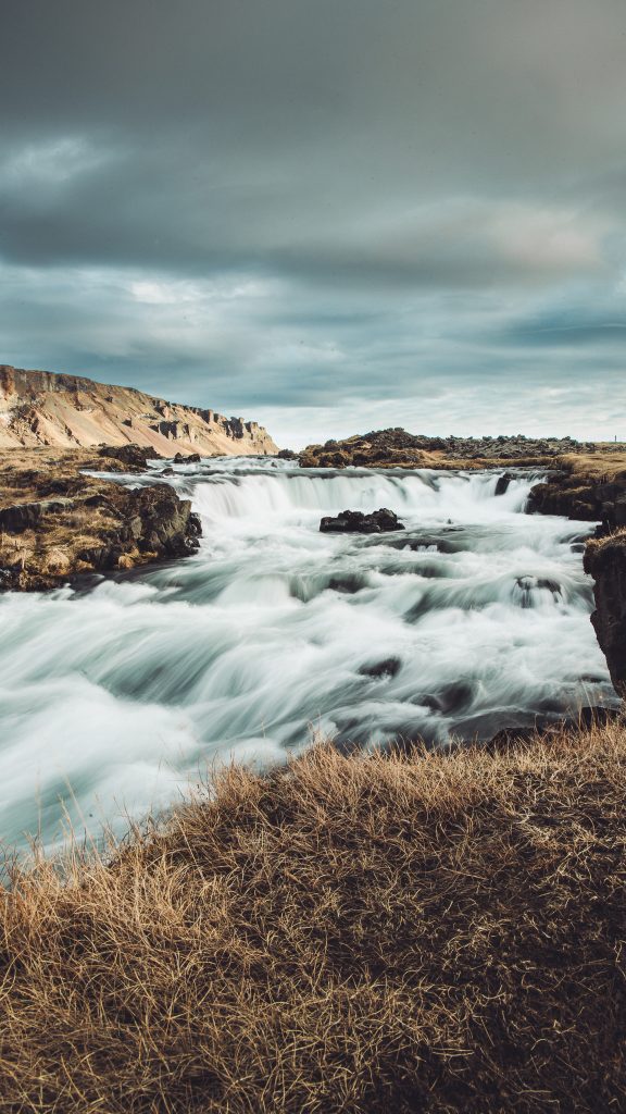 Weite Aufnahme vom Fossálar Wasserfall in Island mit flacher Kaskade, schnell fließendem Wasser, Gras und wolkenverhangenem Himmel