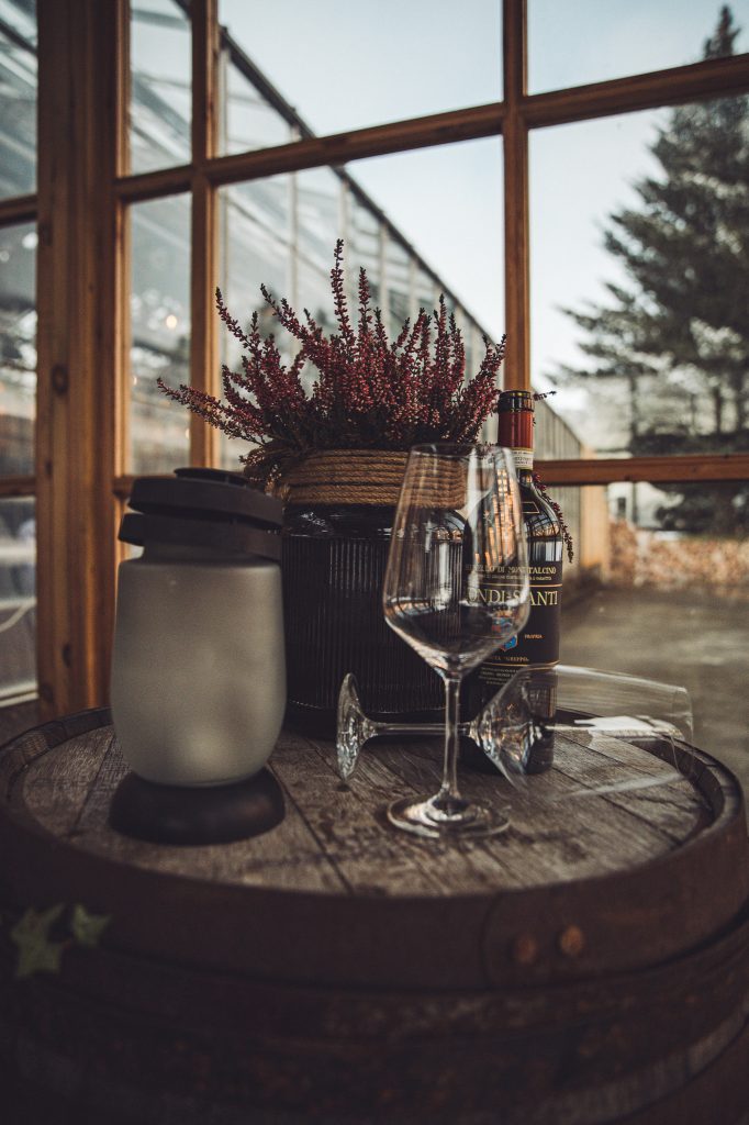 Weinglas und Dekoration auf einem Fass vor Fenstern mit Blick auf das Gewächshaus von Friðheimar