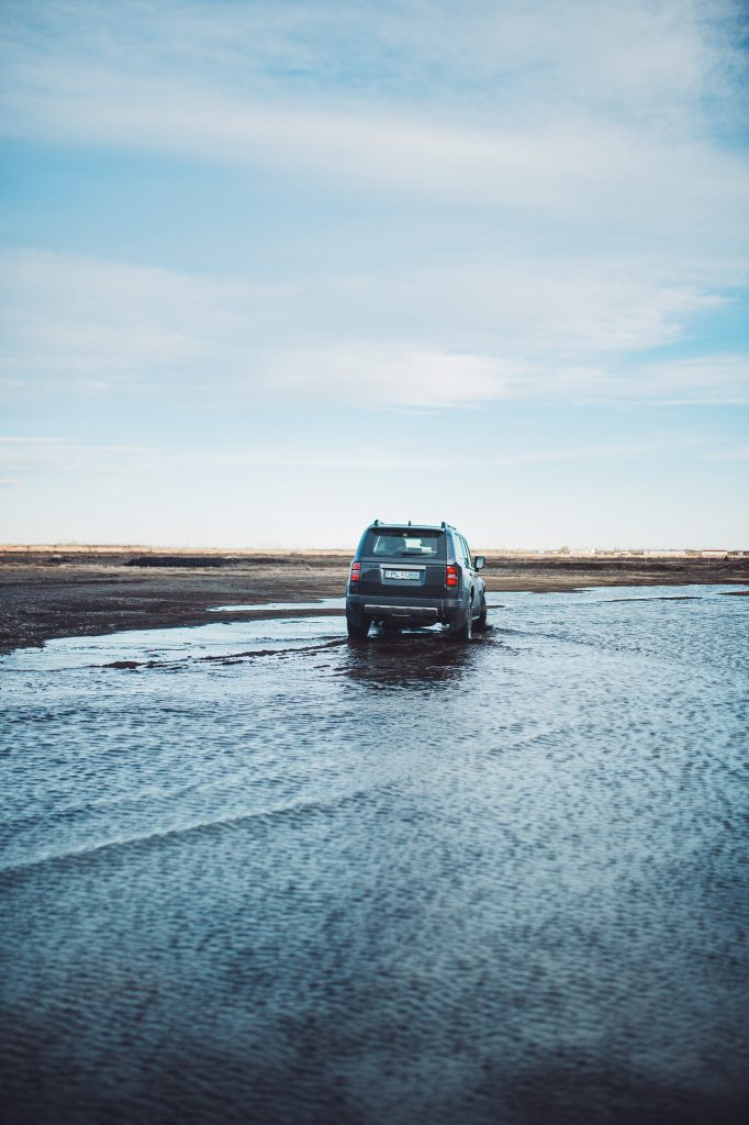 Grauer SUV fährt durch flaches Wasser auf einem teilweise überfluteten Parkplatz nahe dem Stjórnarfoss in Südisland