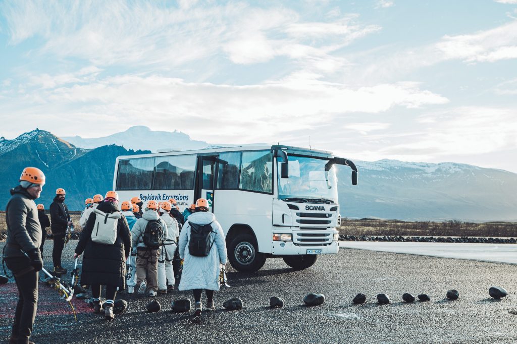 Reisegruppe mit Helmen vor einem weißen Bus in Südisland vor Bergen und Gletscherkulisse auf dem Weg zur Gletscherwanderung in Island