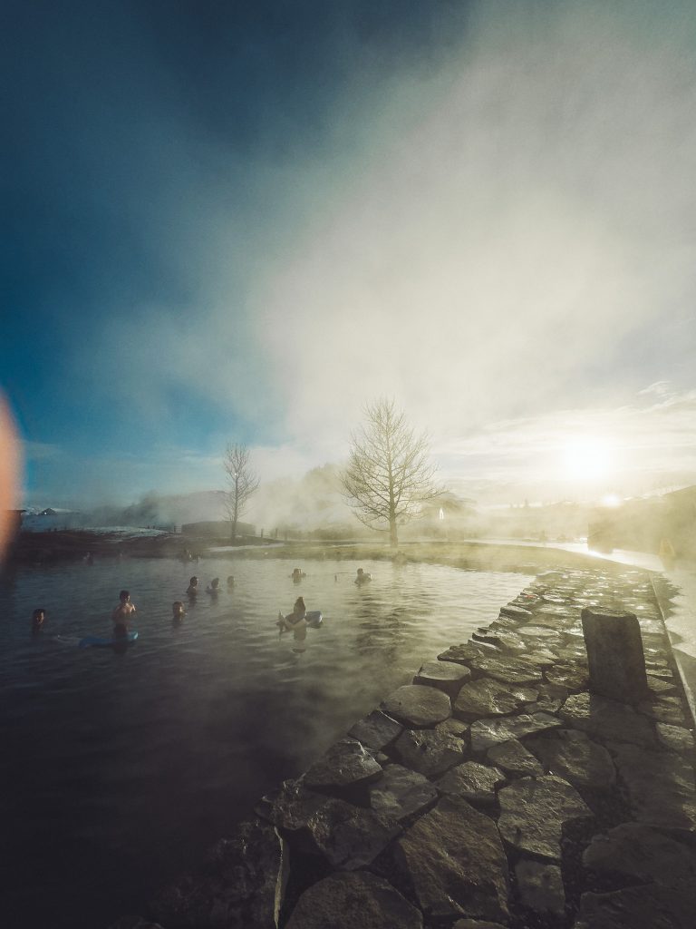 Blick über das warme Becken der Secret Lagoon in Flúðir mit Steinrand, Dampf, Badegästen, kahlen Bäumen und tief stehender Abendsonne.