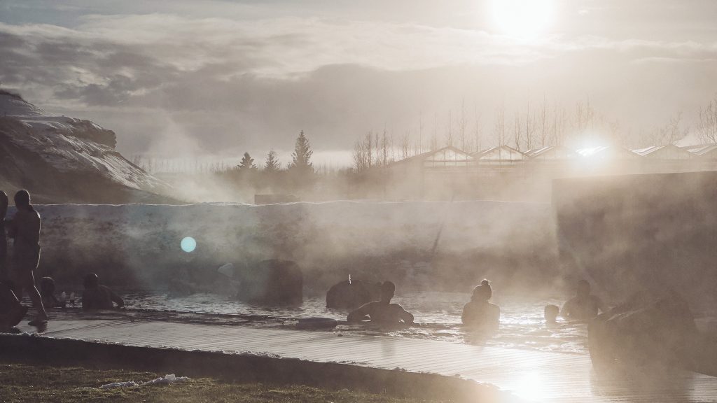 Menschen sitzen im dampfenden Becken der Secret Lagoon in Flúðir, während die tief stehende Sonne durch den Wasserdampf scheint.