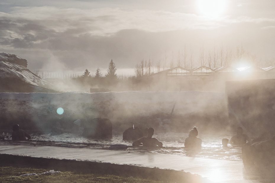 Menschen sitzen im dampfenden Becken der Secret Lagoon in Flúðir, während die tief stehende Sonne durch den Wasserdampf scheint.
