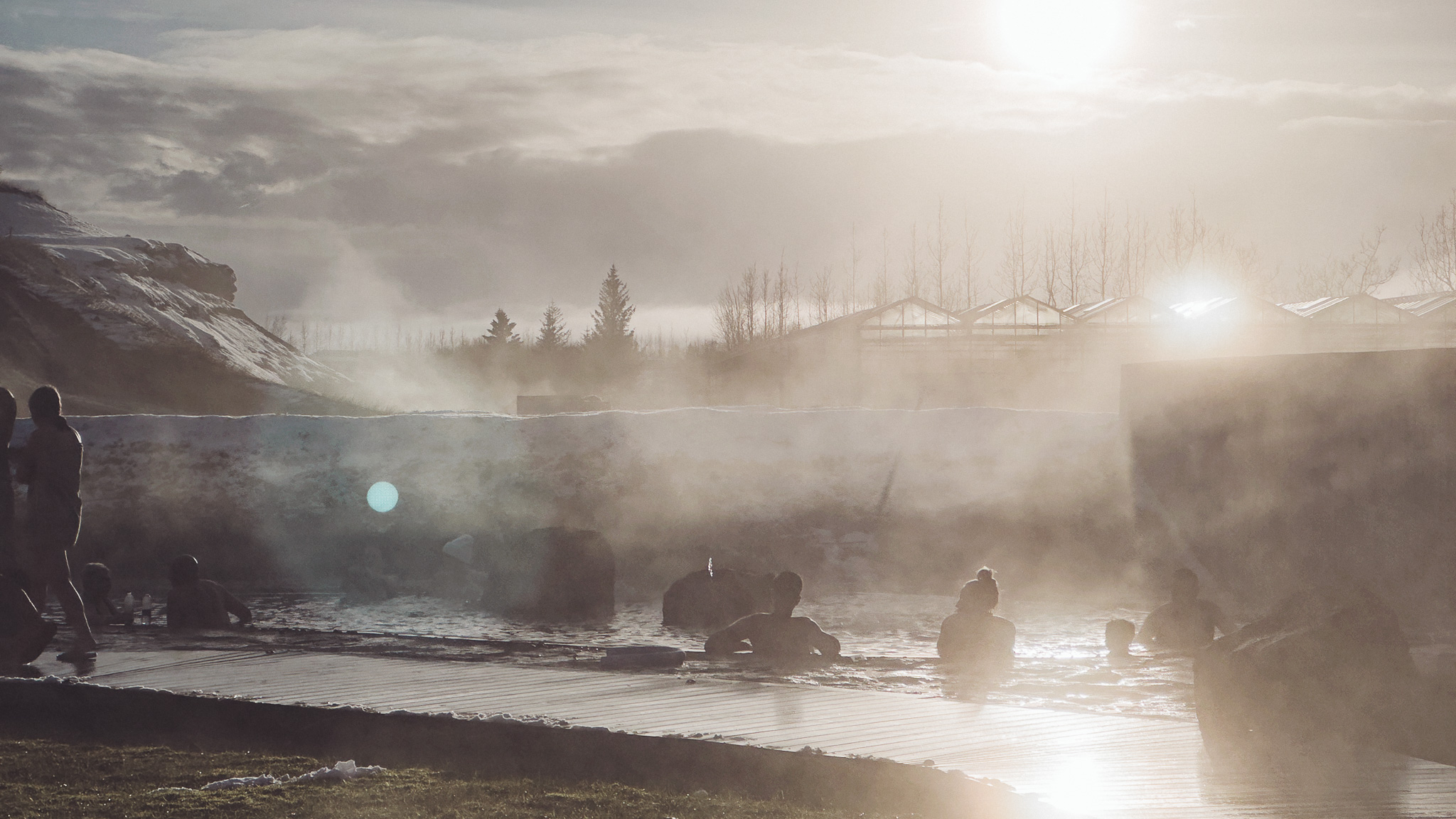 Menschen sitzen im dampfenden Becken der Secret Lagoon in Flúðir, während die tief stehende Sonne durch den Wasserdampf scheint.