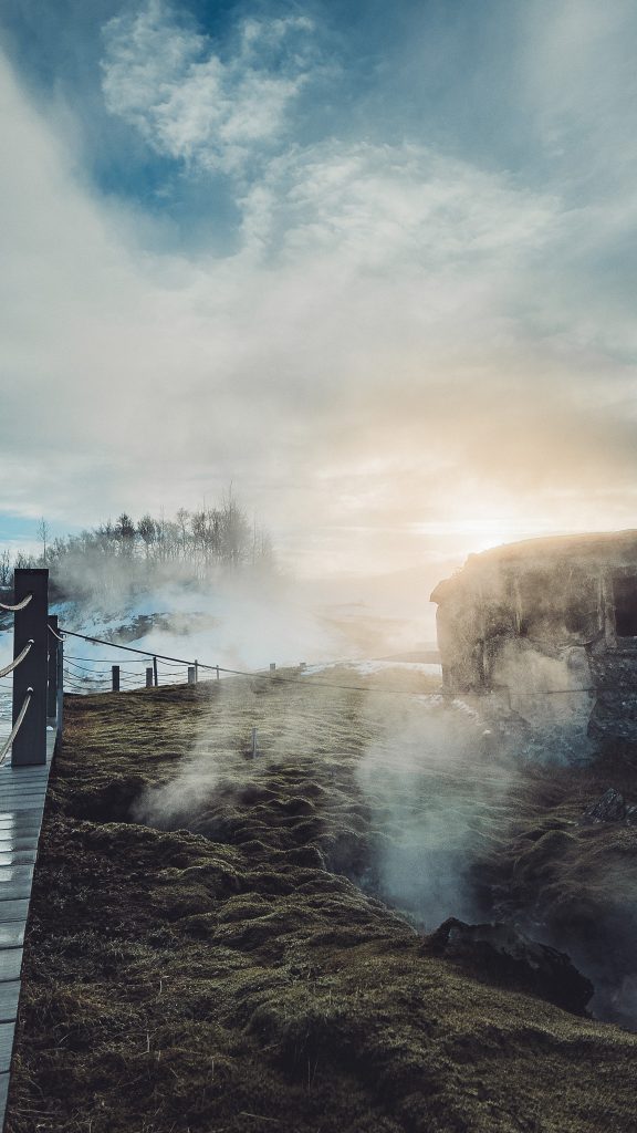 Dampf zieht über moosige heiße Quellen neben dem Holzweg der Secret Lagoon in Flúðir, rechts ragt ein Felsen aus dem Gelände.