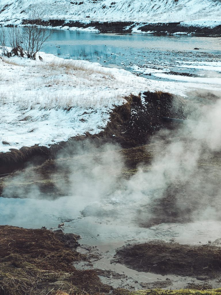 Dampf steigt an einer heißen Quelle nahe des verschneiten Ufers bei der Secret Lagoon in Flúðir auf, dahinter liegt ein kalter Fluss.