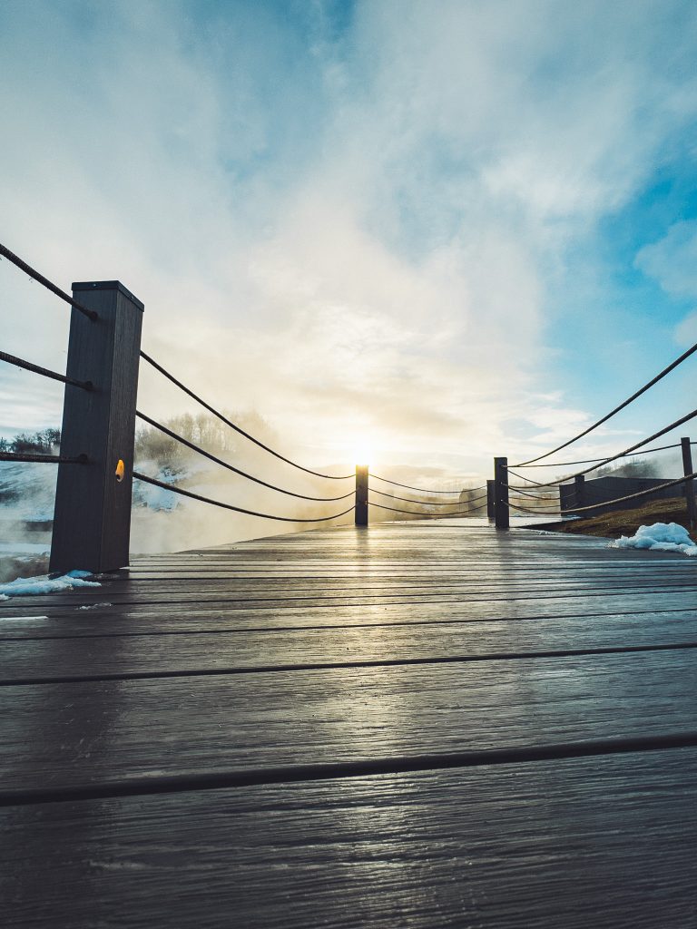 Tiefe Perspektive auf einen nassen Holzsteg der Secret Lagoon in Flúðir mit Seilen, Dampf und tief stehender Sonne am Horizont.