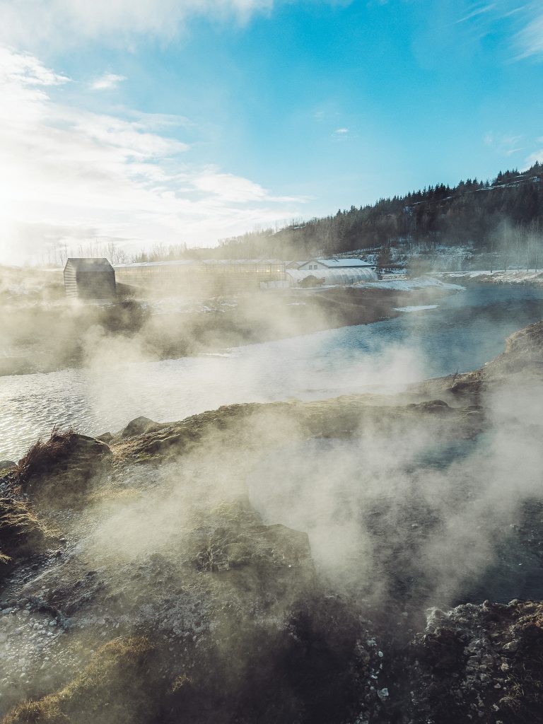 Blick über dampfende heiße Quellen und einen Fluss bei der Secret Lagoon in Flúðir, mit Gebäuden und verschneiter Landschaft im Hintergrund.