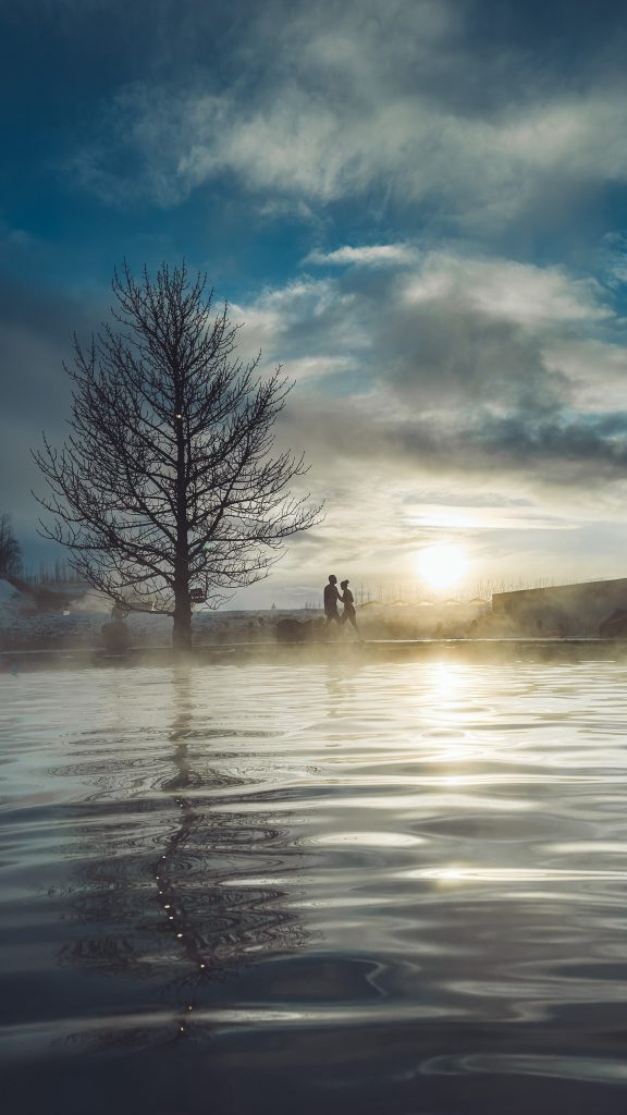 Tiefe Perspektive über die Wasseroberfläche der Secret Lagoon in Flúðir mit kahlem Baum, Spiegelung, Dampf und tief stehender Sonne.