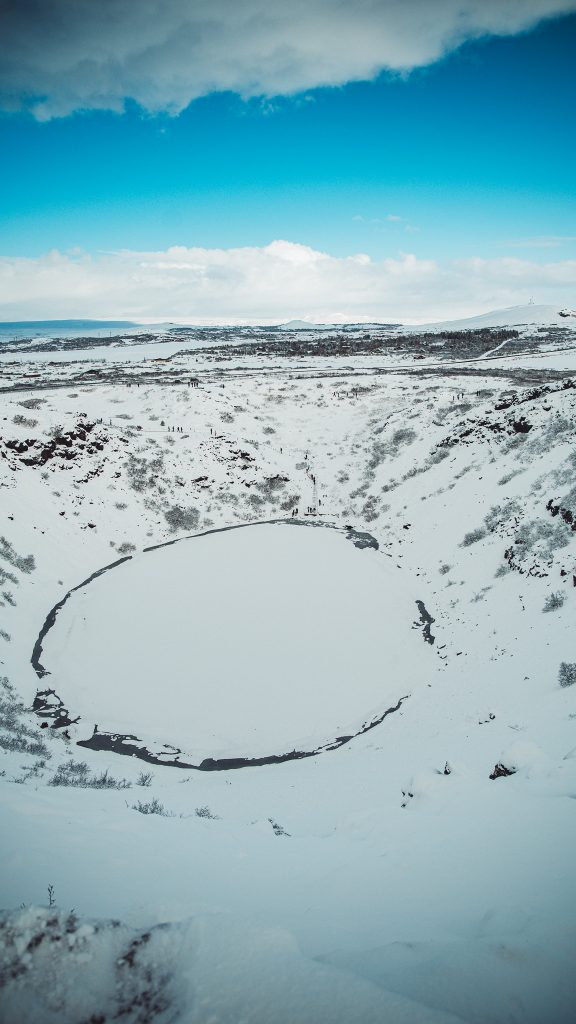 Schneebedeckter Kerið-Krater in Island mit gefrorener Wasserfläche und weiter Winterlandschaft unter blauem Himmel