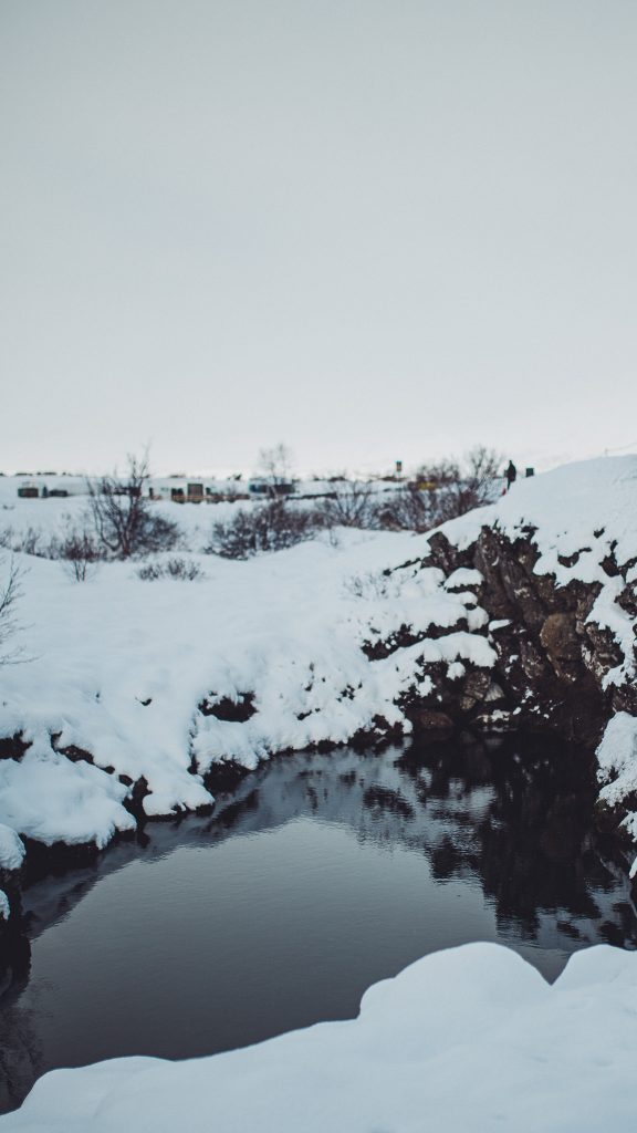Dunkle Wasserfläche zwischen verschneiten Felsen und Ufern im winterlichen Þingvellir-Nationalpark
