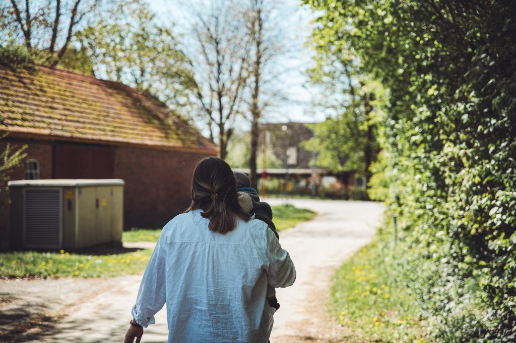 Frau trägt Kind auf dem Hofweg am Biohof Hümme zwischen Backsteingebäude und grüner Hecke