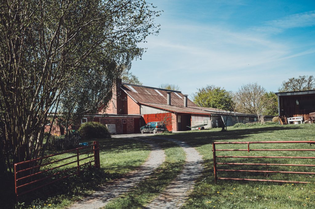 Hofzufahrt am Biohof Hümme mit Scheune, rotem Gatter, Wiese und altem Hofgebäude