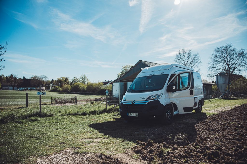 Weißer Camper auf dem Stellplatz am Lindenhof Broosch mit Acker, Wiese und Hofgebäuden im Hintergrund.