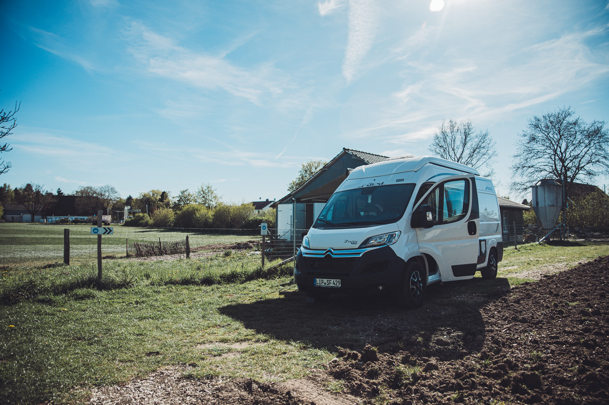 Weißer Camper auf dem Stellplatz am Lindenhof Broosch mit Acker, Wiese und Hofgebäuden im Hintergrund.