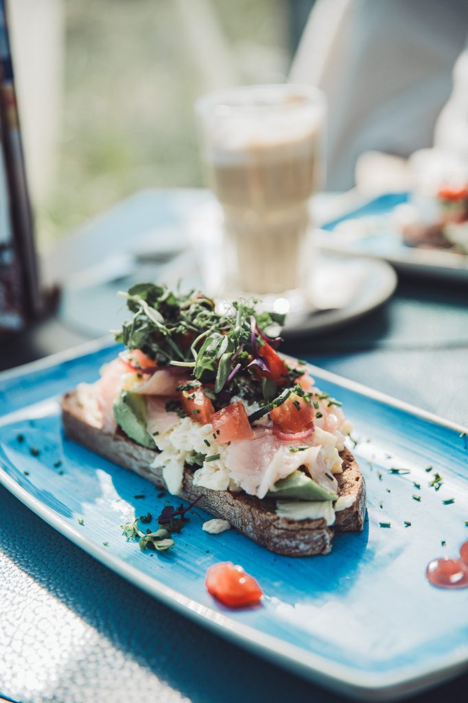 Sand und Stulle mit Fisch, Avocado und Rührei auf blauem Teller mit Kaffee im Hintergrund