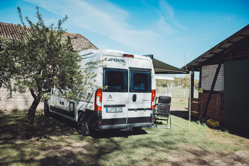 Rear view of a white Pössl camper van beside a brick shed, with a leafy tree in the foreground and a clear blue sky in the background.