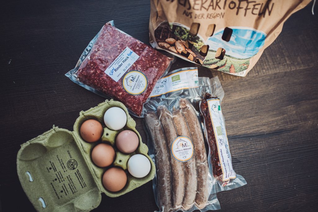 Assorted groceries on a dark table: egg carton with brown eggs, sausages in plastic, a loaf of bread, and a paper bag behind them.
