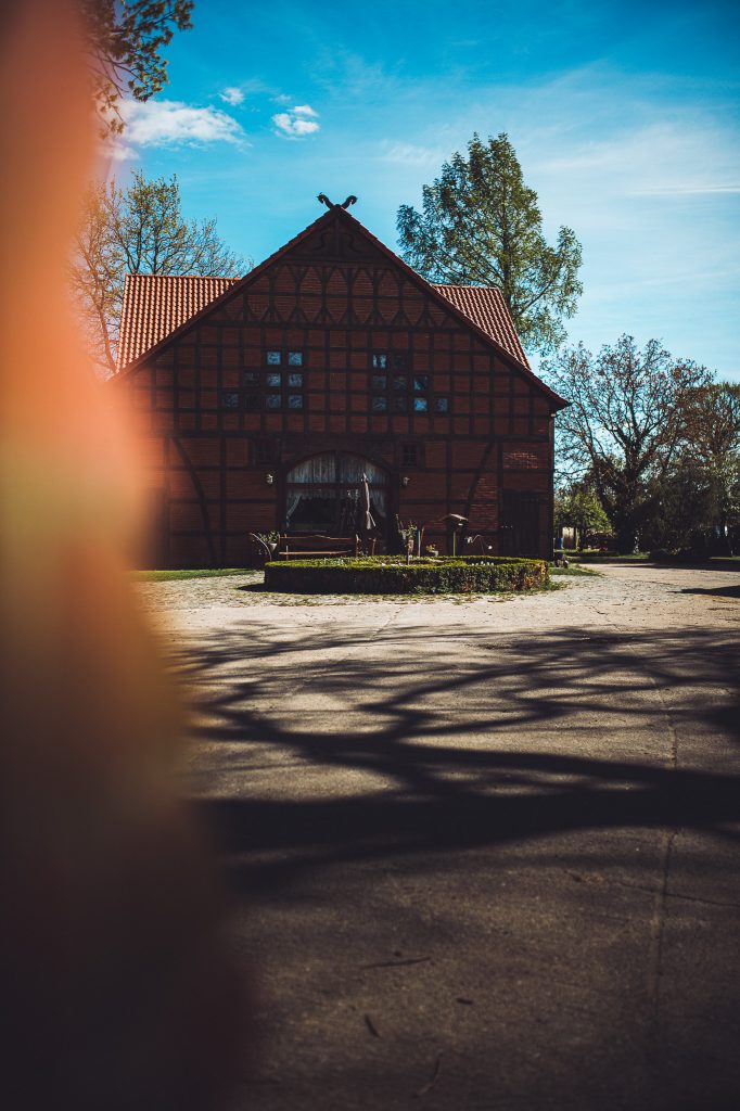 Timber-framed brick house with a red-tiled roof in a sunny courtyard, seen with a blurred foreground element on the left.