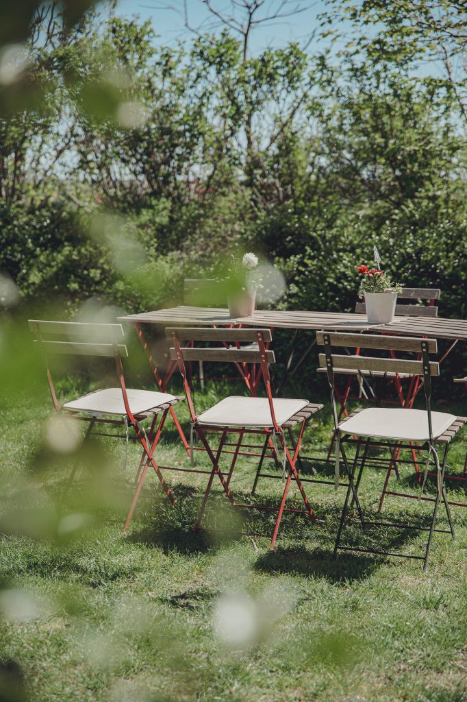 Outdoor dining setup with wooden tables and red-legged chairs on a grassy lawn, potted flowers on the tables, and a green hedge in the background.