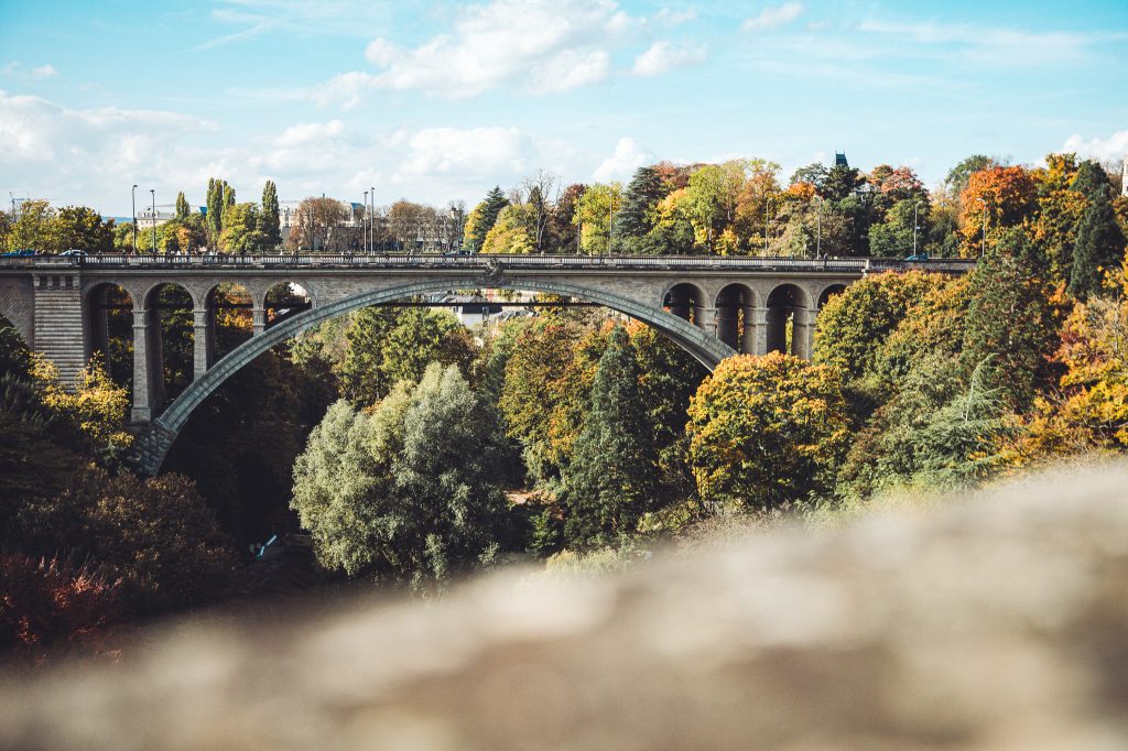 Blick auf eine große Steinbrücke über dem grünen Pétrusse-Tal in Luxemburg Stadt mit Herbstbäumen und blauem Himmel.